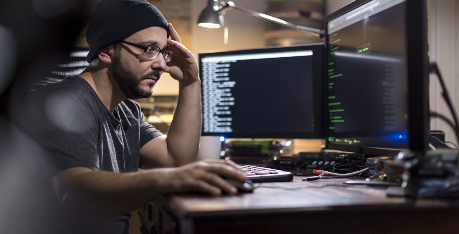 Man with black stocking cap sitting at computer in fairly dark room, representing a programmer or hacker