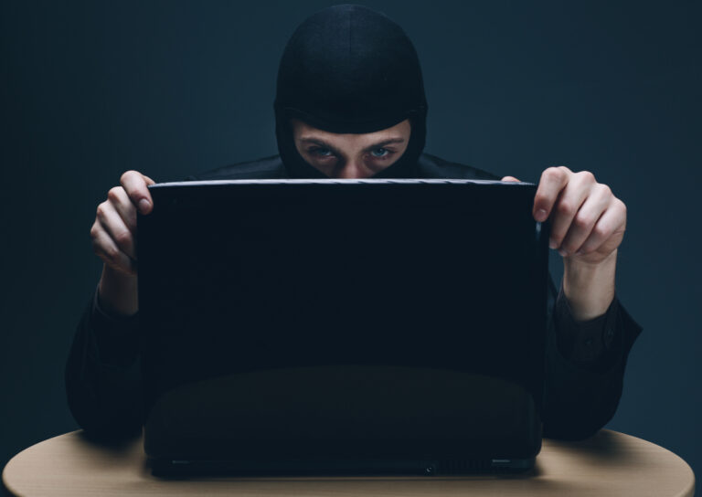 Photo of man in black mask behind open laptop; representing identity theft, cybercrime