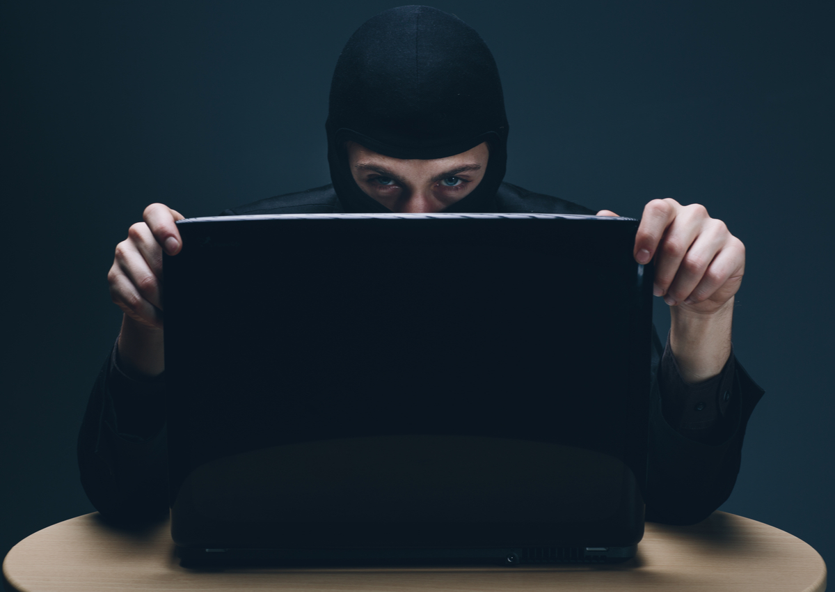 Photo of man in black mask behind open laptop; representing identity theft, cybercrime